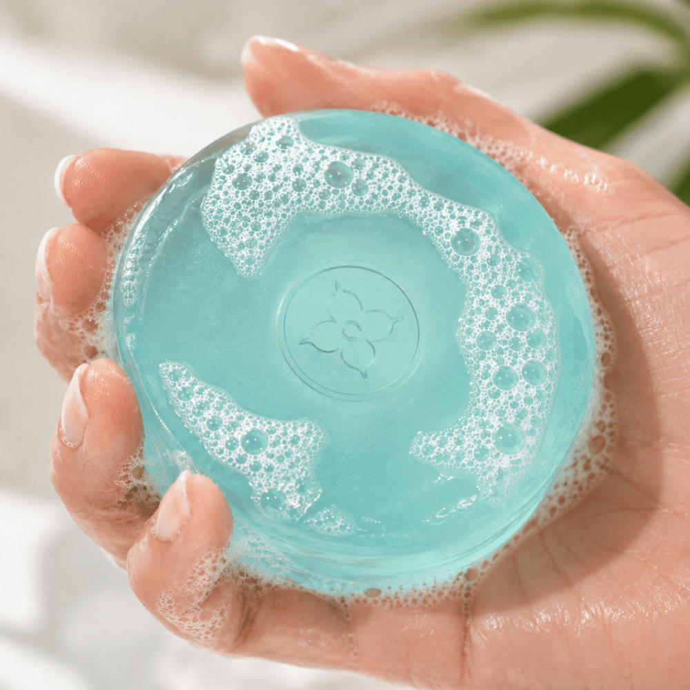 Mint - A hand holding a round, translucent aqua soap bar covered in white foam, with a subtle floral embossed design in the center.